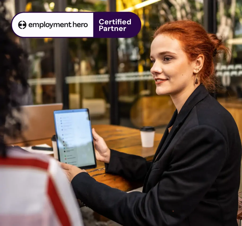 Young woman with red hair in a black blazer holding a tablet, engaged in conversation at a cafe. "Employment Hero Certified Partner" text in corner.