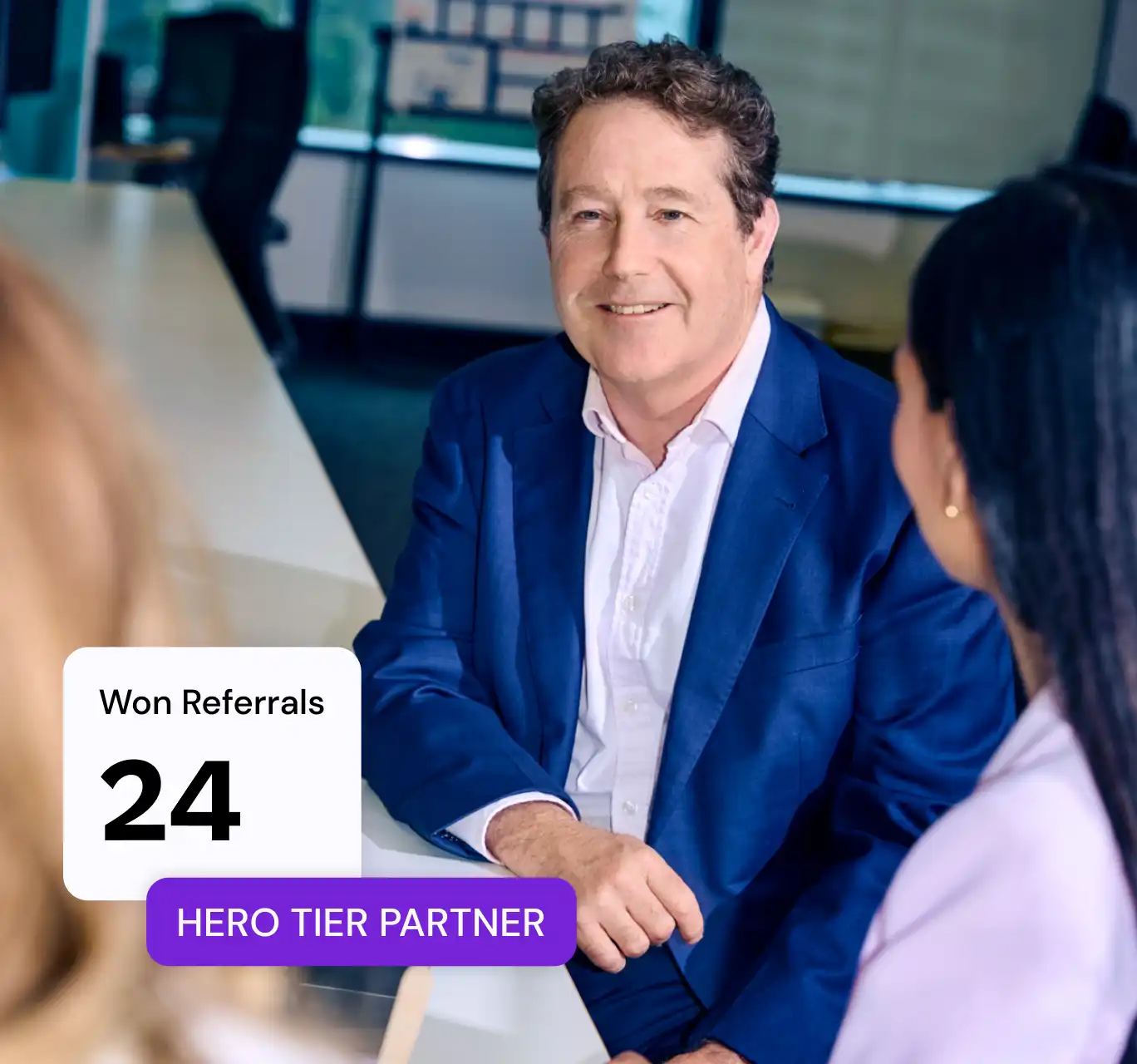 A man in a blue suit smiles at two colleagues in an office setting. A graphic shows "Won Referrals: 24" and "Hero Tier Partner," indicating success.