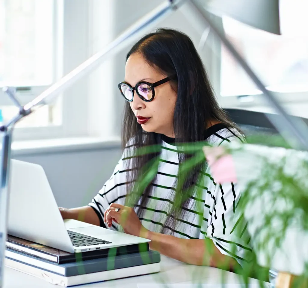 A woman in glasses and a striped shirt works intently on a laptop in a bright office, surrounded by plants, creating a focused yet serene atmosphere.