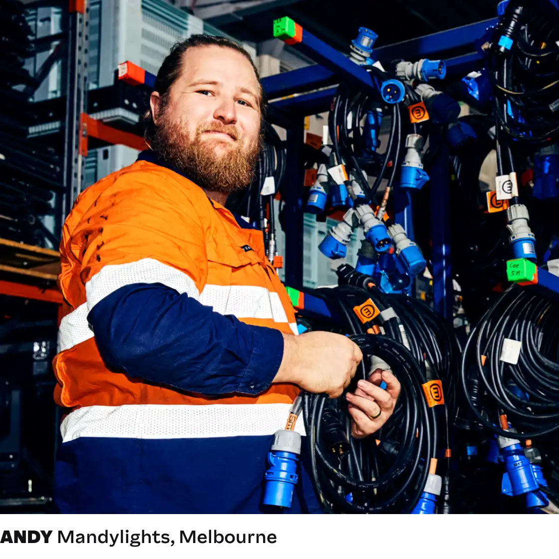 A person in a high-visibility orange work shirt stands confidently among numerous coiled cables with blue connectors, suggesting an organized workspace.