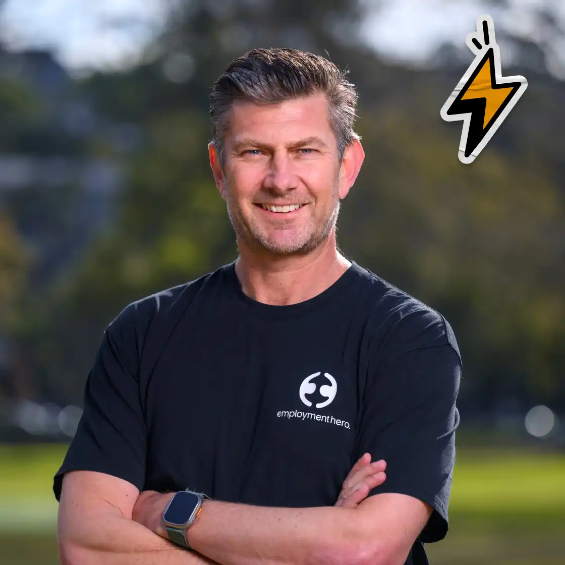 Smiling man in a black "Employment Hero" shirt stands outdoors with arms crossed. Background is a blurred park. A lightning bolt icon is on the top right.