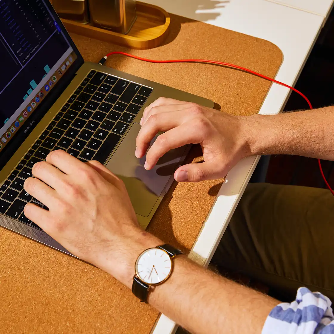Two hands typing on a laptop keyboard on a cork mat, with a watch on one wrist. A red cable and a warm sunlit tone create a focused work atmosphere.