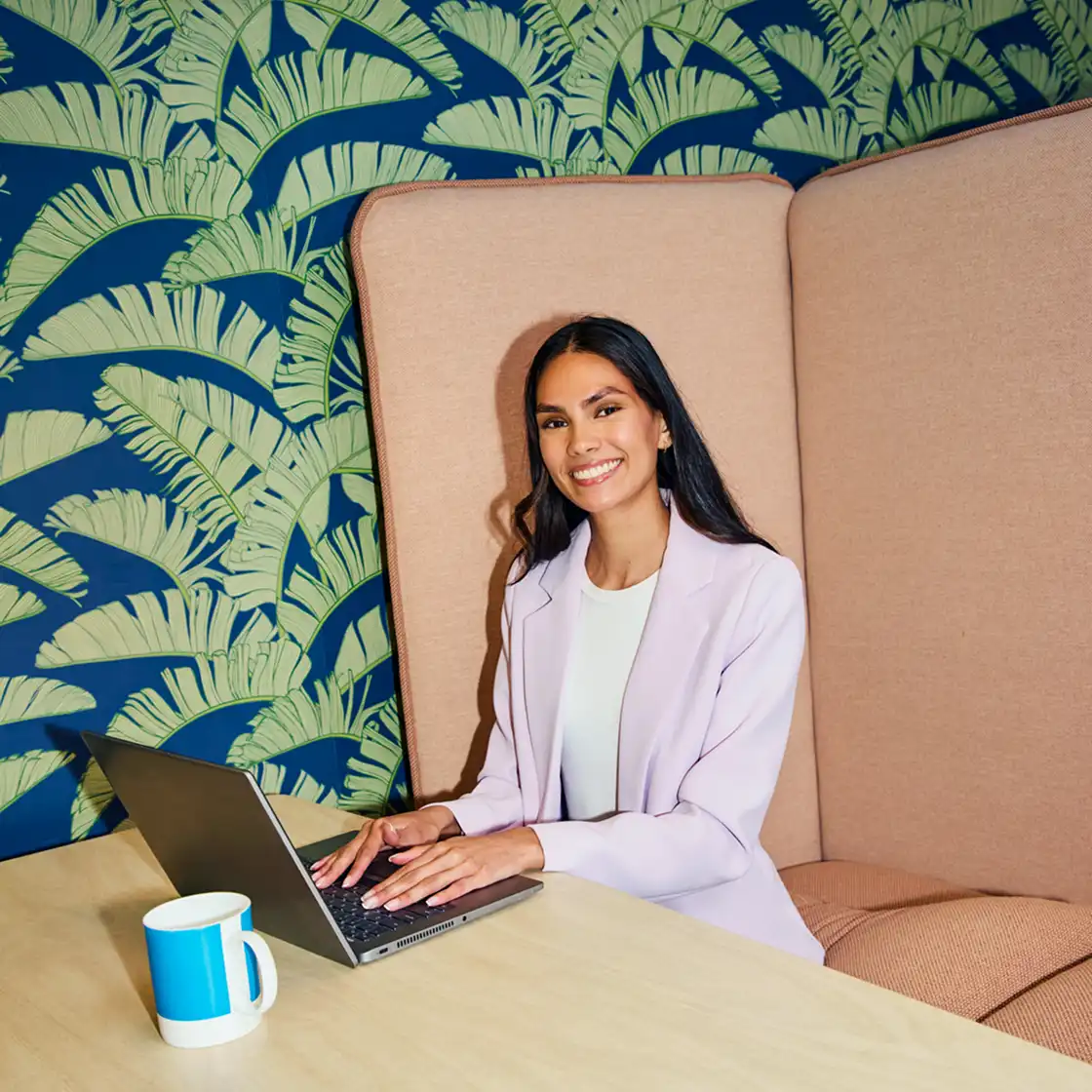 Smiling woman in a light blazer uses a laptop at a wooden table, with a blue mug beside her. Background features a vibrant leaf-patterned wallpaper.