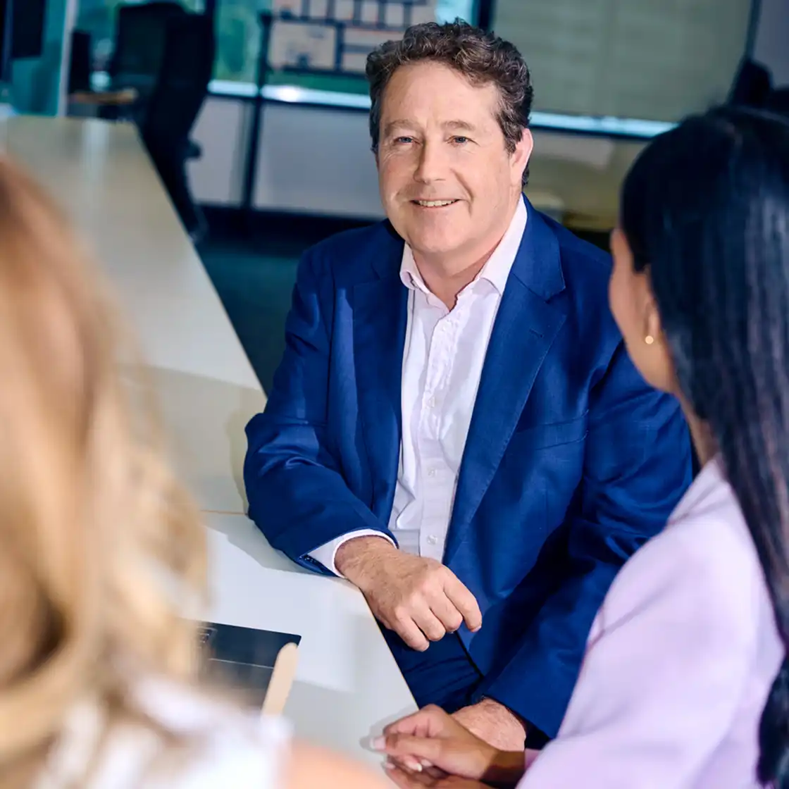Man in a blue suit, smiling and engaged in conversation with two women at an office table. The atmosphere is professional and friendly.