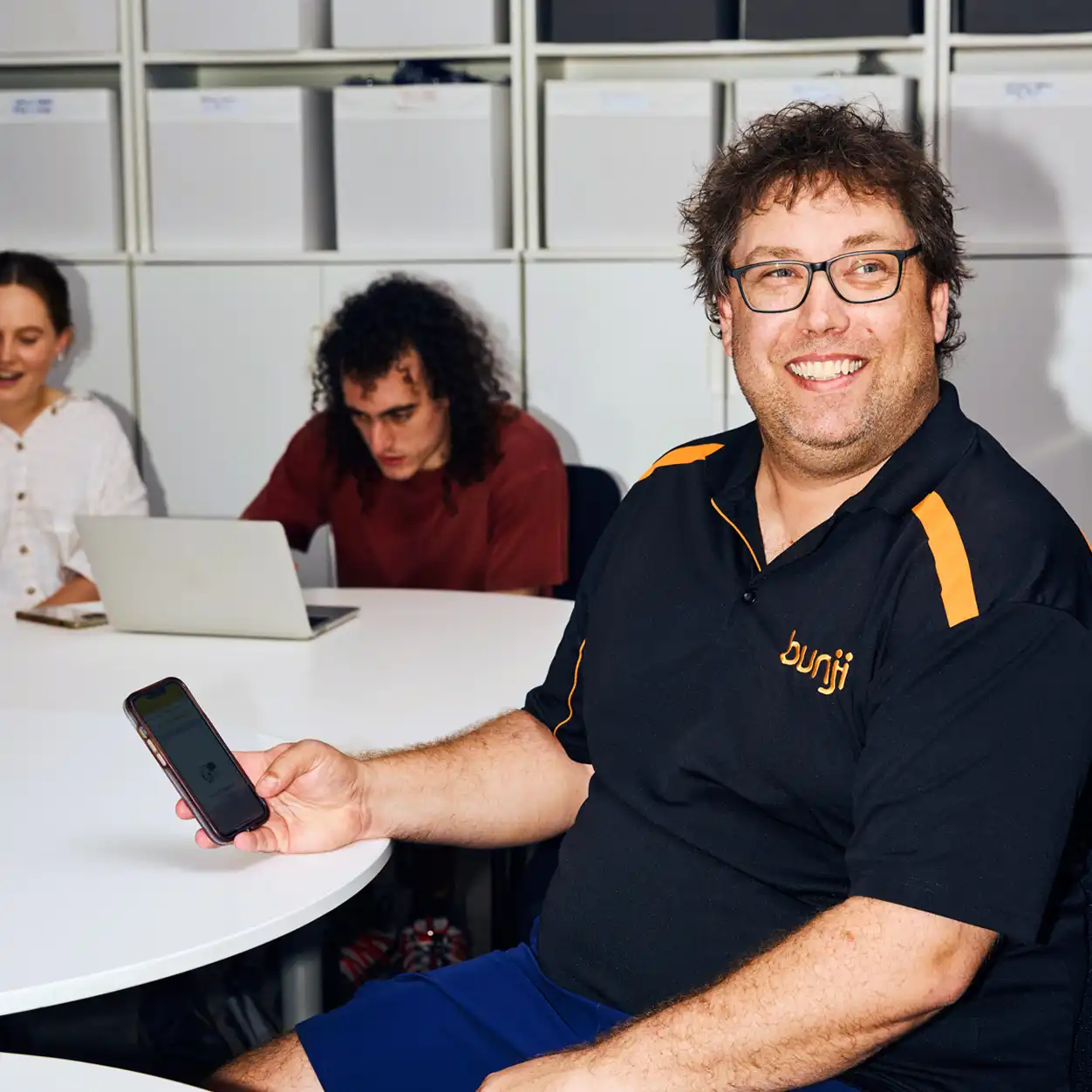 A smiling man in a black polo shirt holds a smartphone at a round table. Two people in the background focus on a laptop, suggesting a collaborative setting.