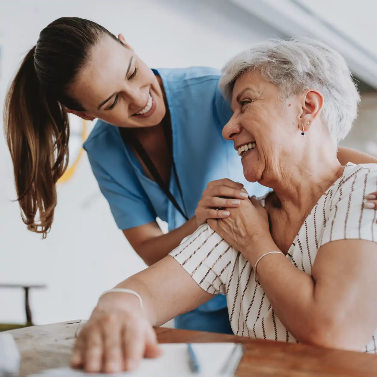 A caregiver in blue scrubs smiles warmly at an elderly woman with short gray hair, who is also smiling. They are sharing a moment of joy and comfort.
