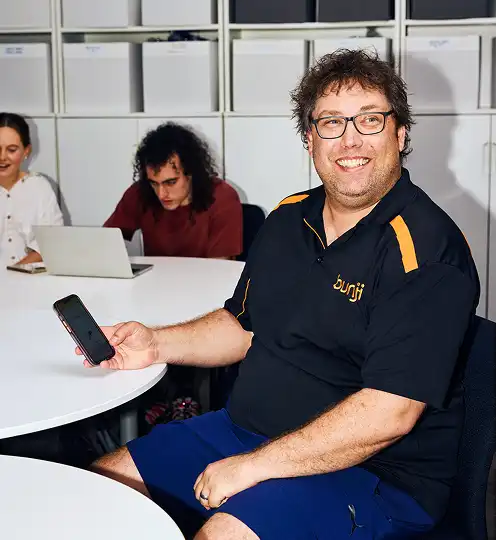 A smiling man wearing glasses and a black shirt with yellow accents holds a phone, seated at a table. Two people work on laptops in the background. The atmosphere is casual and collaborative.