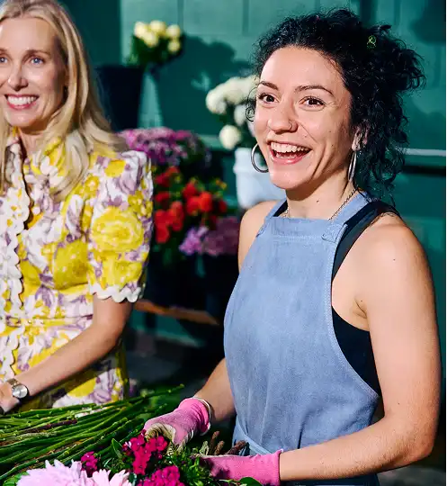 Two women standing in a flower shop, smiling as they arrange bouquets. One wears a yellow floral dress, the other a blue apron and pink gloves.