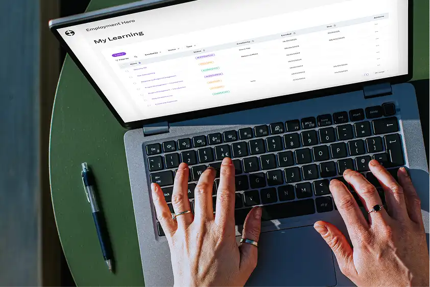 Hands typing on a laptop showing "My Learning" portal with course list, atop a green table beside a pen, indicating productivity and focus.