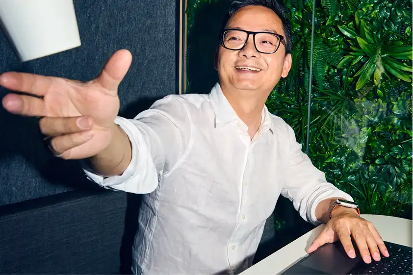 A man in glasses, wearing a white shirt, joyfully tosses a cup indoors. He sits by a laptop, with vibrant green plants in the background, creating a lively atmosphere.