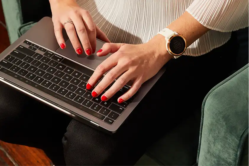 Hands with red nail polish type on a laptop keyboard. A smartwatch adorns one wrist, and the person wears a white, pleated blouse, conveying focus and professionalism.