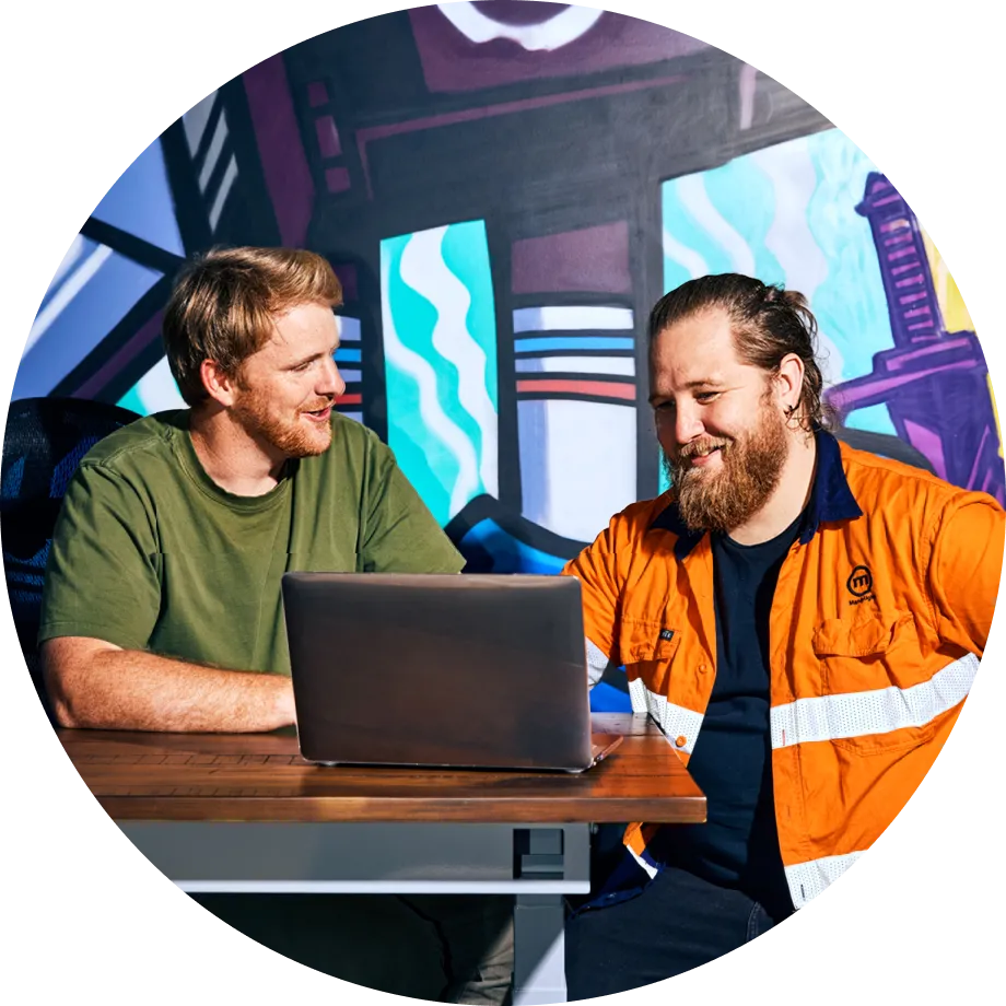 Two men are sitting at a desk, smiling and discussing something while looking at a laptop. One man is wearing a green t-shirt, and the other is wearing an orange high-visibility work shirt with white reflective stripes.