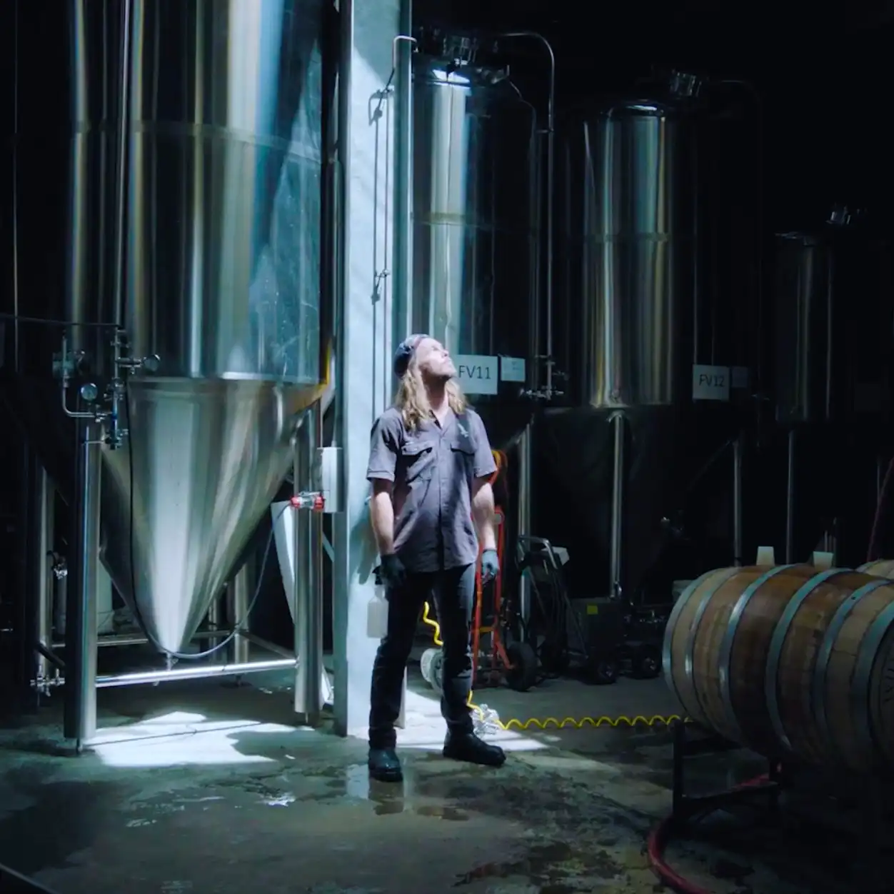 A person stands in a brewery, gazing up at large stainless steel fermentation tanks. The space is dimly lit, creating a focused and industrious atmosphere.