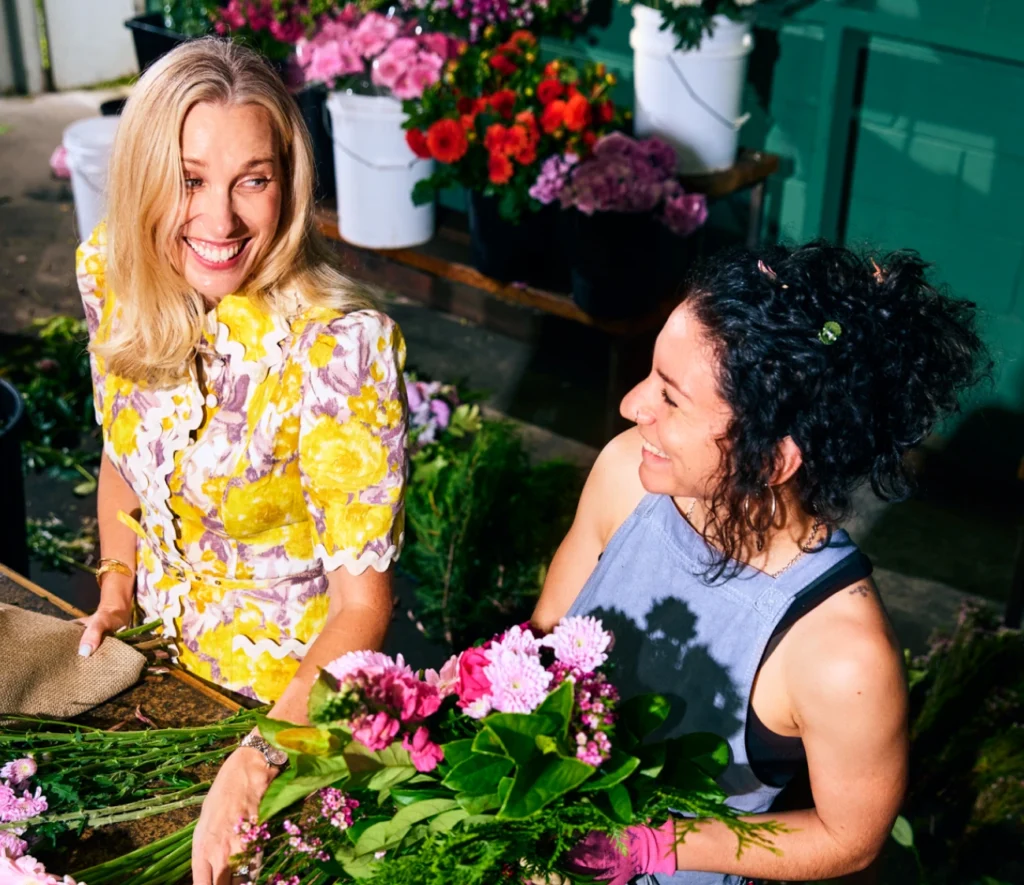Two women smiling and arranging fresh flowers at a florist shop.