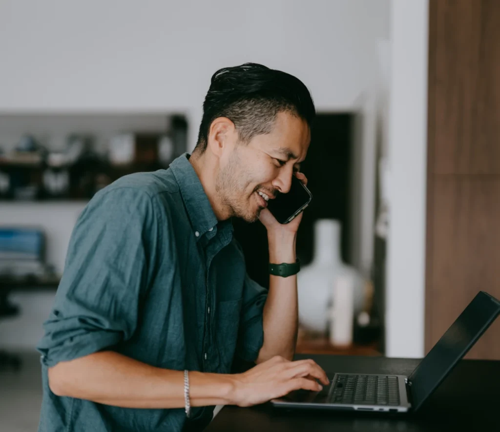 Man smiling while talking on the phone and typing on a laptop in a home office.