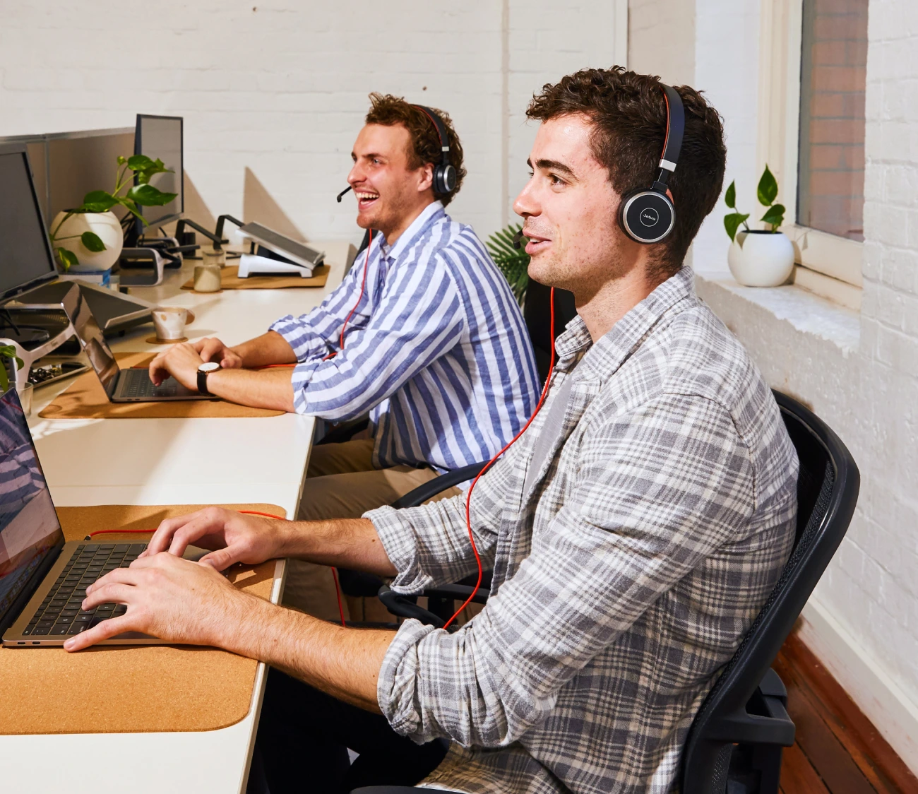 Two men wearing headsets smiling while working at laptops in a customer support office.
