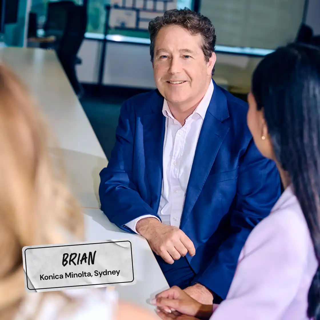 A man in a blue suit sits at a white table in a modern office, smiling at two colleagues. Bright, professional atmosphere with large windows.