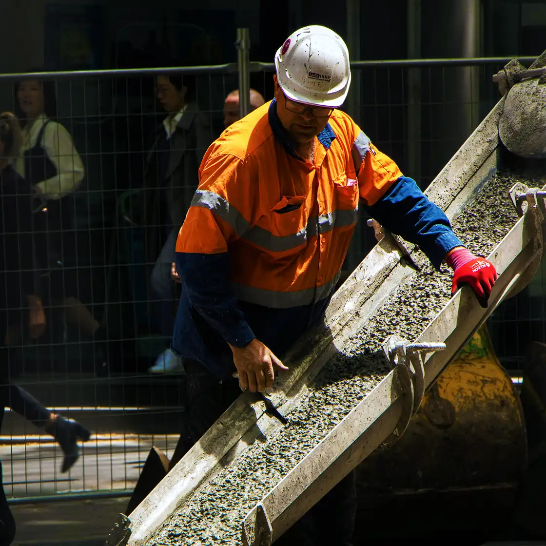 A construction worker in an orange safety jacket and white helmet spreads concrete on a chute. Behind a fence, blurred pedestrians convey urban bustle.