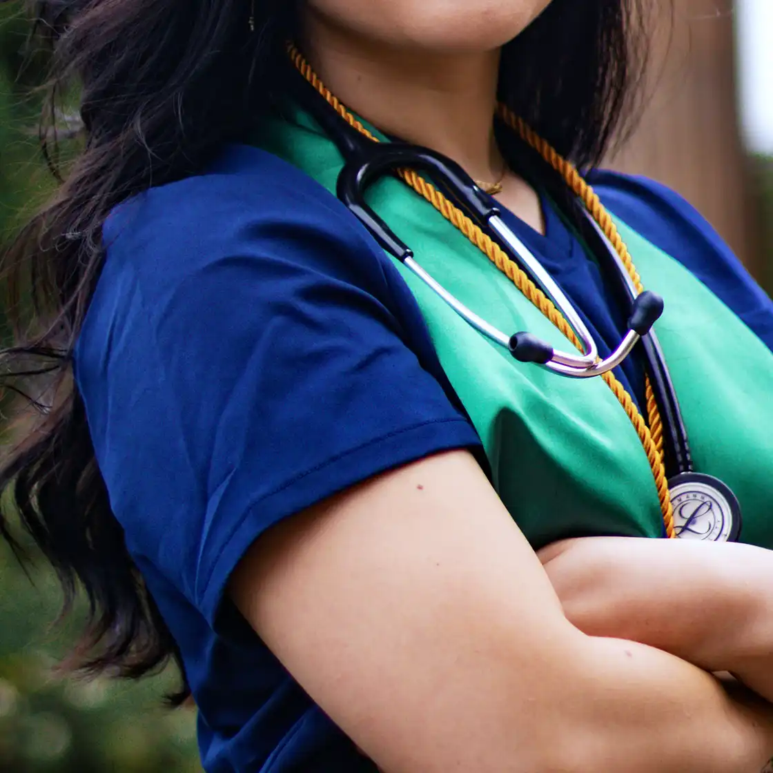 Young woman in a dark blue top with a green sash and gold rope, arms crossed. A stethoscope hangs from her neck, suggesting a medical student.