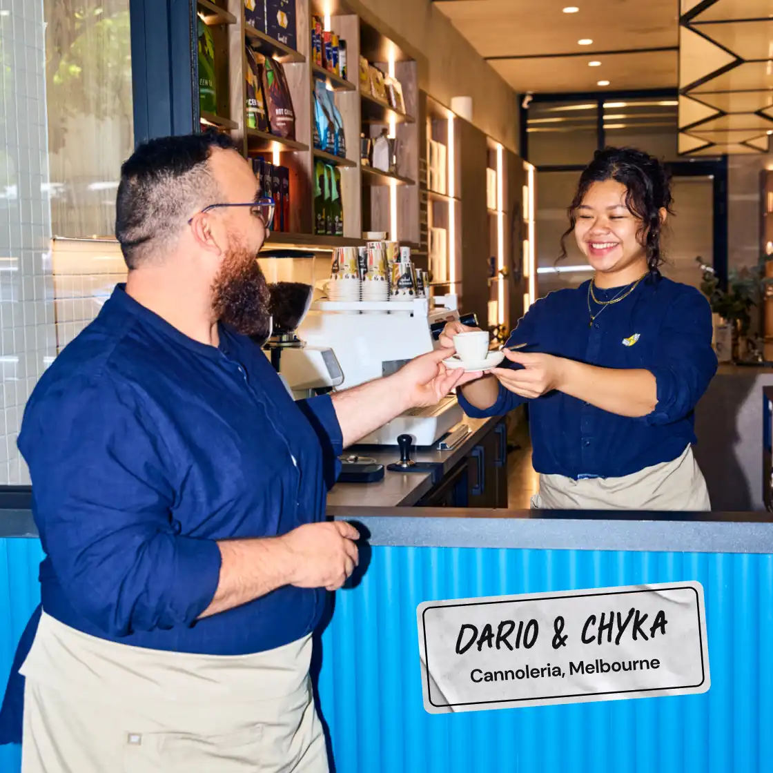 A woman in a café smiles as she hands a coffee cup to a bearded man. They wear matching blue shirts and aprons. Sign reads "Dario & Chyka, Cannoleria, Melbourne."