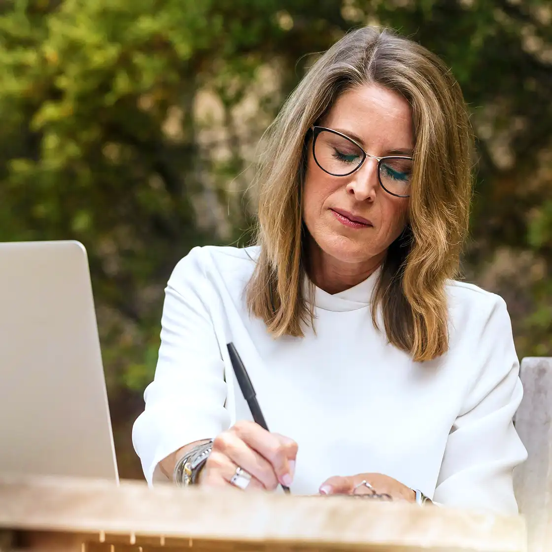 A woman with glasses writing in a notebook outdoors. She is focused and wearing a white blouse, with a laptop beside her. Greenery is in the background.