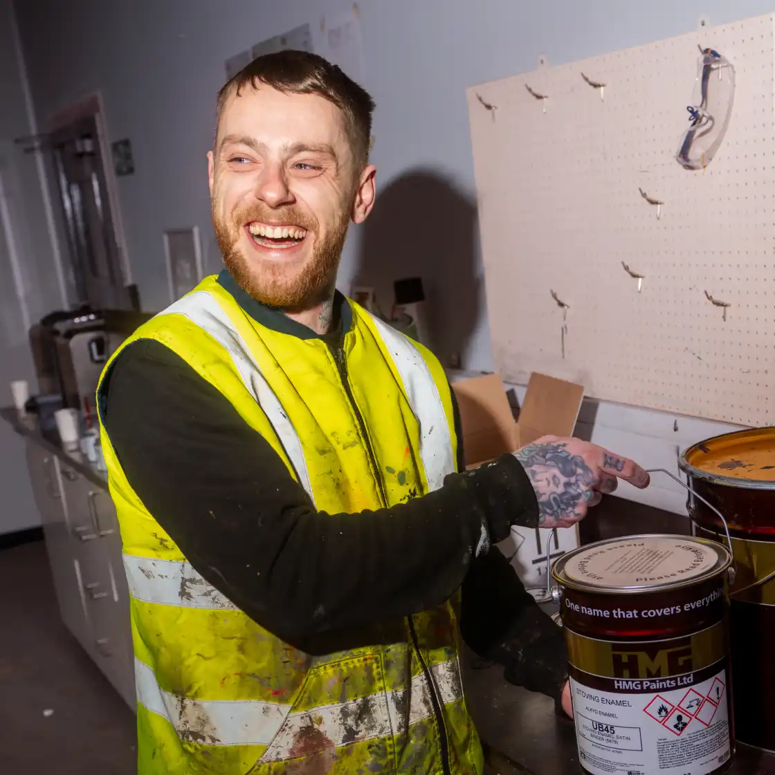 Man in a high-visibility vest with tattoos on his hand smiles while pointing at a paint can in a workshop. The mood is cheerful and lighthearted.