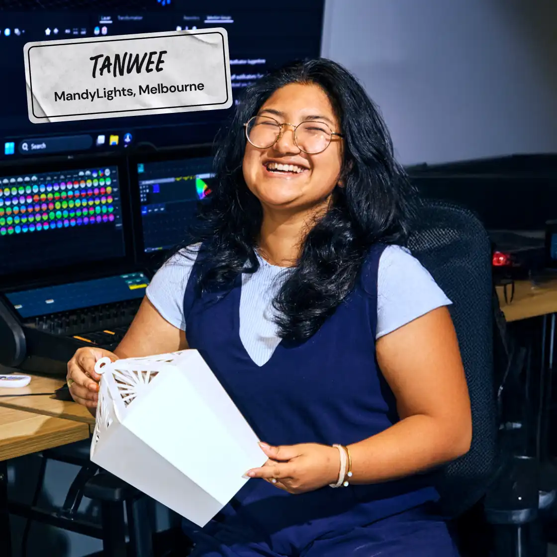 Smiling person with long hair and glasses, holding a white geometric object inside a studio. In the background, computer screens display colorful graphics.