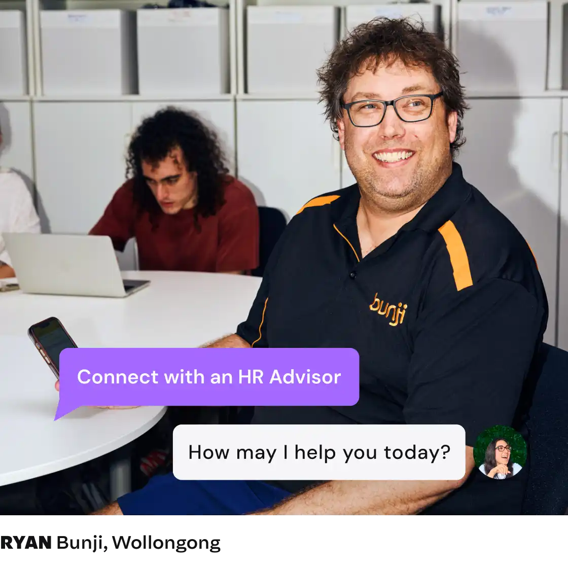 A smiling man in glasses wearing a black shirt with orange accents sits at a table, holding a phone. Text bubbles read "Connect with an HR Advisor" and "How may I help you today?" Another person with curly hair works on a laptop in the background, creating a collaborative and professional tone.