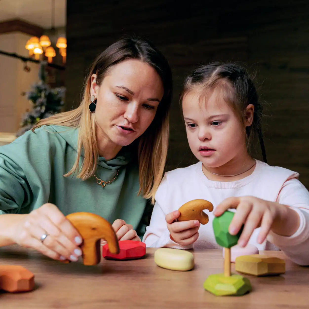 A woman and a young girl with braided hair are focused on playing with colorful wooden toys at a table, creating a warm and educational atmosphere.